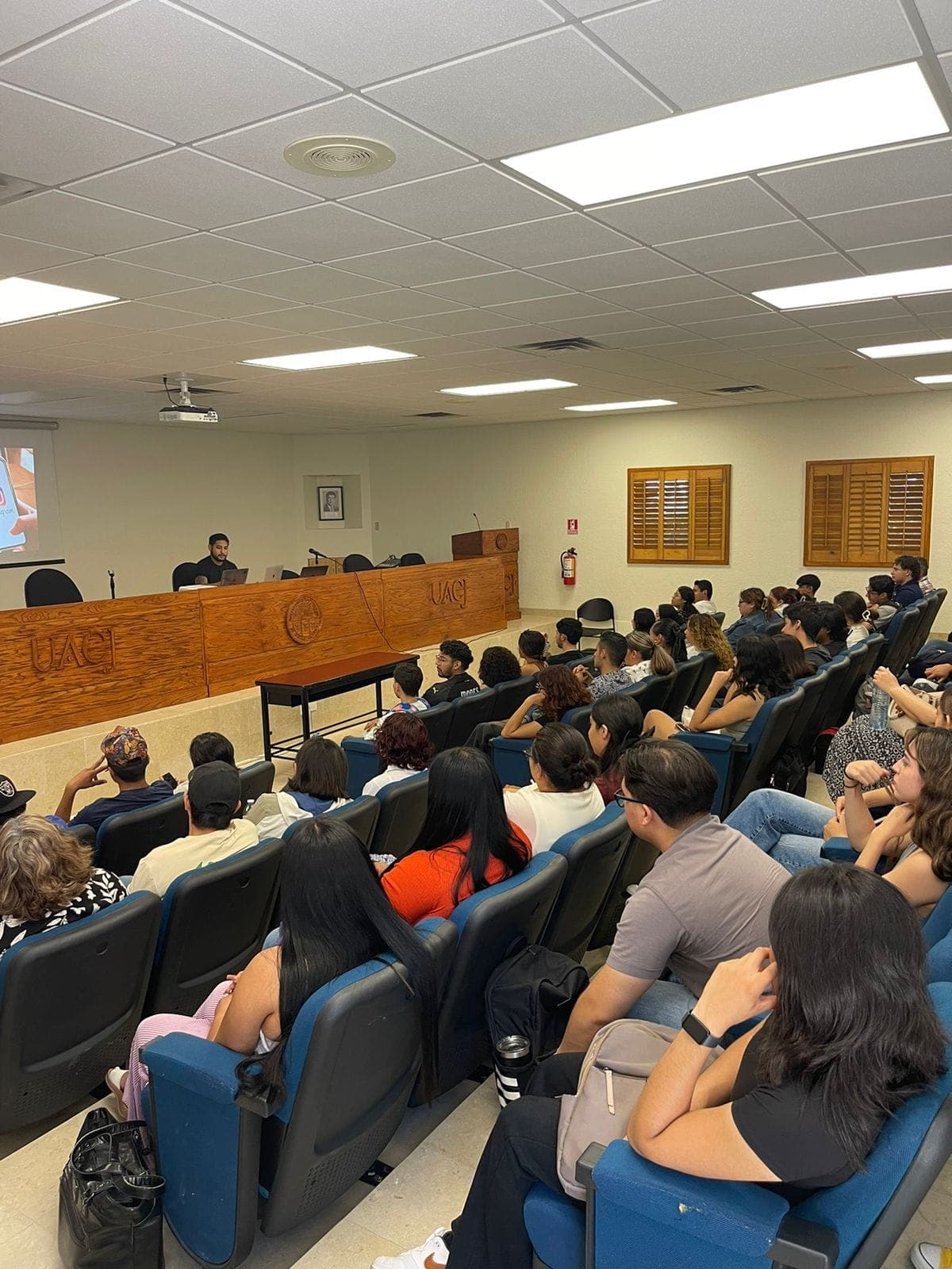 Packed UACJ auditorium seen from the back, with Ramses presenting at the front.